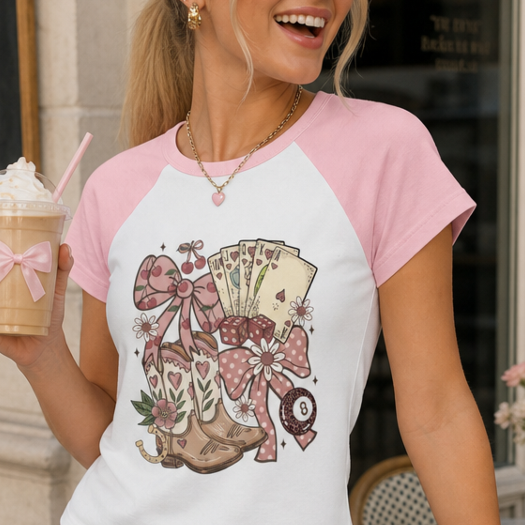 Close-up of a pink coquette baby tee with lucky casino graphic including playing cards, dice, bows and horseshoes.